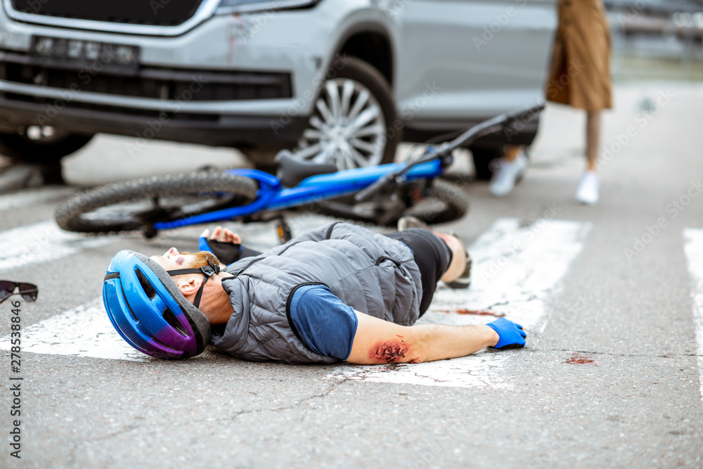 Obraz premium Road accident with injured cyclist lying on the pedestrian crossing near the broken bicycle and worried woman driver and car on the background