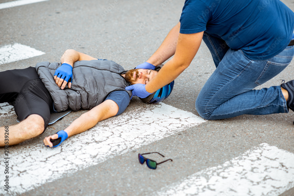 Road accident with injured cyclist on the pedestrian crossing with passerby pedestrian providing first aid fixing men's head Road accident with injured cyclist on the pedestrian crossing with passerby pedestrian providing first aid fixing men's head