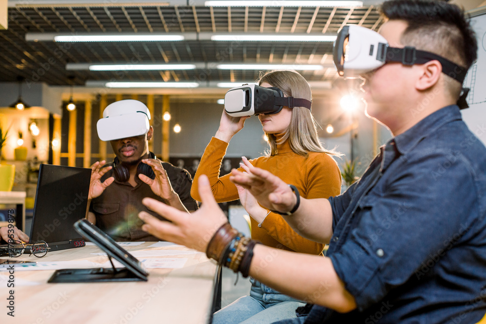 Young friends having fun together with virtual reality headset sitting around the table. Happy people using modern vr goggle glasses. New trends and technology concept Young friends having fun together with virtual reality headset sitting around the table. Happy people using modern vr goggle glasses. New trends and technology concept