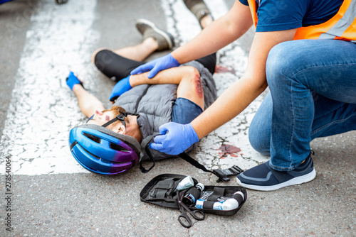 Road accident with injured cyclist lying on the pedestrian crossing near the bicycle and car, male driver providing first aid