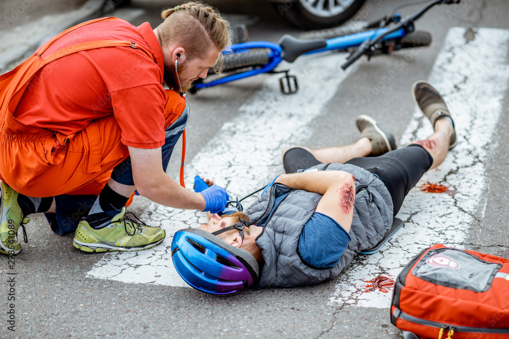 Medic applying first aid to the injured cyclist lying on the pedestrian crossing after the road accident, doctor measuring pressure Medic applying first aid to the injured cyclist lying on the pedestrian crossing after the road accident, doctor measuring pressure