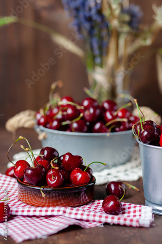 Composition with ripe cherries in metal bowls and clay brown plate. Vintage napkins as decor. Dacha style  countryside  cozy and cute  grandmother s kitchen. Dry flowers in vase on background