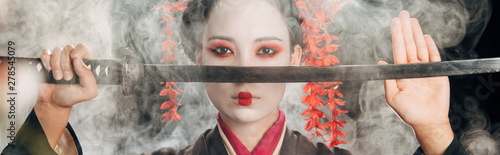 selective focus of beautiful geisha in black kimono holding katana in smoke, panoramic shot