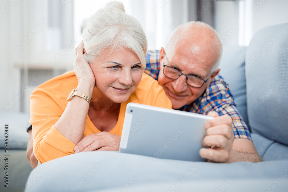Happy senior couple looking at tablet lying on sofa at home
