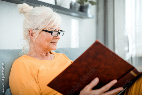 Cheerful senior female holding family photo album sitting on sofa at home