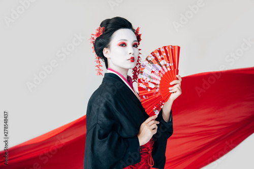 Fotografia beautiful geisha in black kimono with hand fan and red cloth on background isola