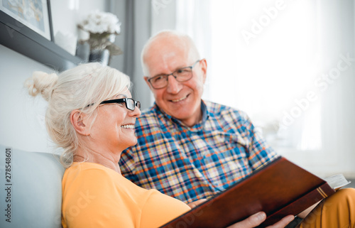 Cheerful senior couple holding family photo album sitting on sofa at home