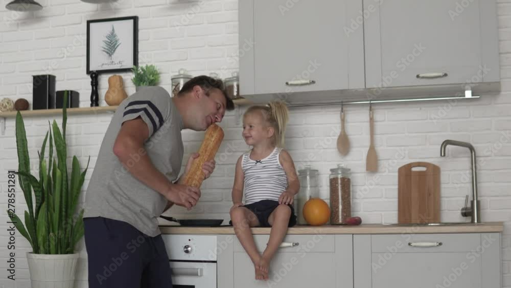 Morning dad and his little daughter in the kitchen. A man plays with a child while he is preparing breakfast, he sings in a loaf of bread, as if it were a microphone and laughs Morning dad and his little daughter in the kitchen. A man plays with a child while he is preparing breakfast, he sings in a loaf of bread, as if it were a microphone and laughs