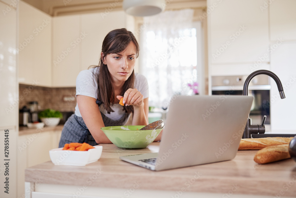 Pretty Caucasian brunette in apron leaning on kitchen counter and looking at laptop and following recipe for dinner. Woman holding carrot in hand. Pretty Caucasian brunette in apron leaning on kitchen counter and looking at laptop and following recipe for dinner. Woman holding carrot in hand.