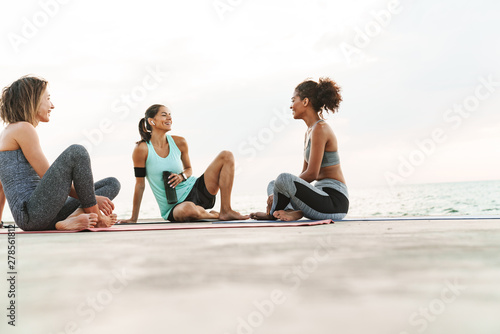 Photo of happy multiethnic sportswomen smiling and talking while sitting on yoga mats by seaside in morning