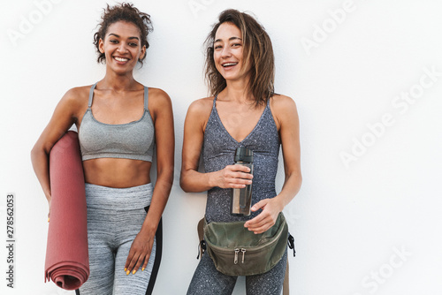 Photo of two young multiethnic sportswomen smiling and holding yoga mat while standing over white wall outdoors photo