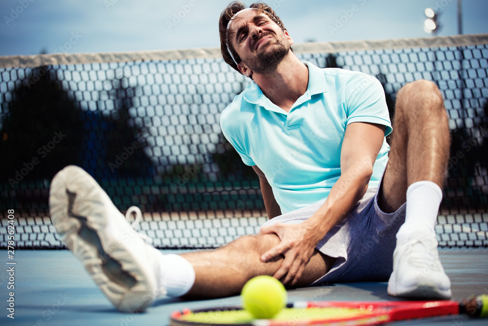 Sports injury. Young tennis player touching his knee while sitting on the tennis court - obrazy, fototapety, plakaty 