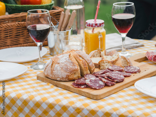 Party in the countryside. Table detail of a picnic decorated outdoors in summer with red wine in glasses, Italian sausage, homemade bread on a wooden cutting board on checkered tablecloth
