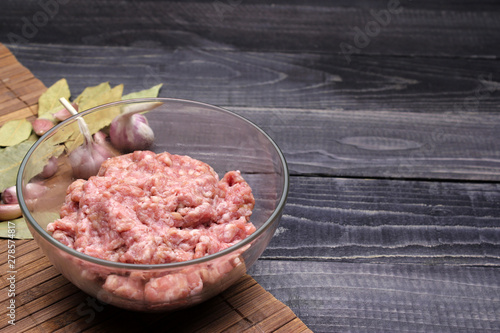 raw minced meat in glass bowl on brown amboo napkin with garlic and spices on black wooden table top surface. Burger. 