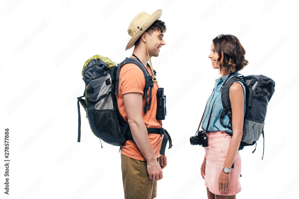 two young, smiling tourists with backpacks looking at each other isolated on white