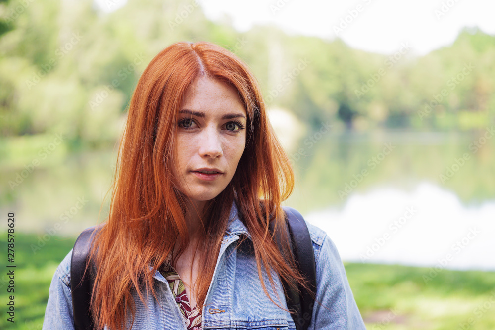 authentic young woman wearing denim jacket and backpack outdoors on a hike - real person standing by lake authentic young woman wearing denim jacket and backpack outdoors on a hike - real person standing by lake