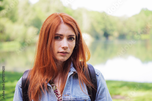 authentic young woman wearing denim jacket and backpack outdoors on a hike - real person standing by lake
