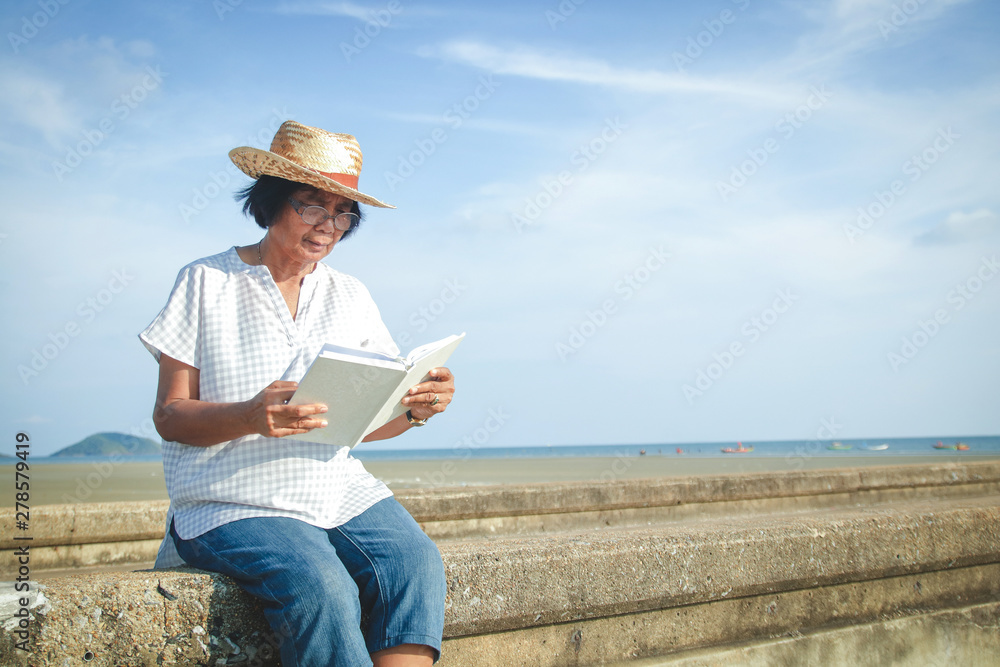 An elderly Asian woman reading a book at the concrete bridge by the sea to relax and breathe fresh air.