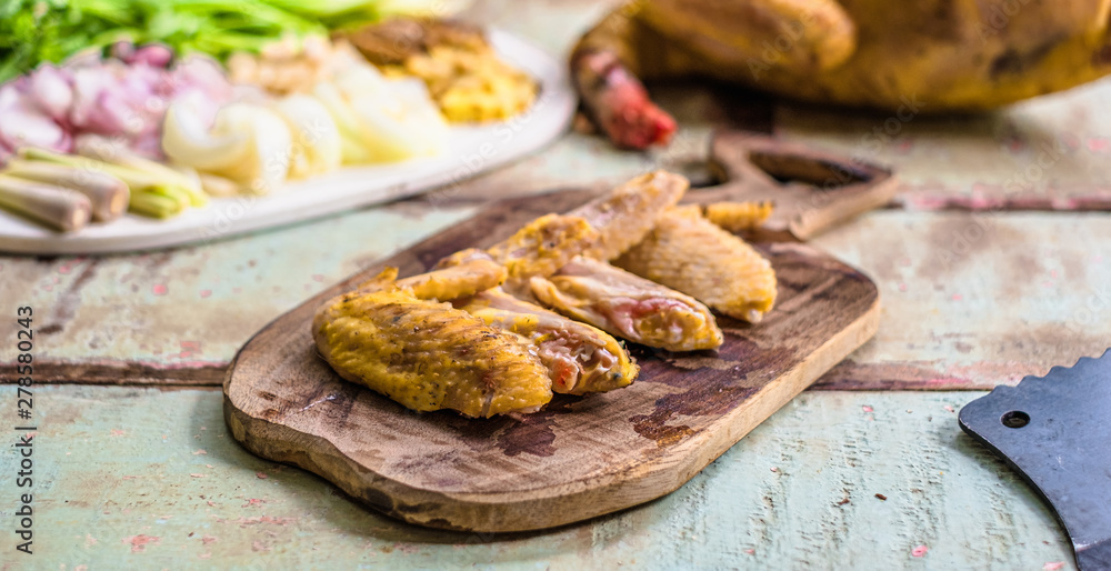 Fresh raw chicken wings on a cutting board on wooden old plank or table with blur Thai herbs, whole chicken and black knife. Chicken with Thai herbs prepared for cooking soup.