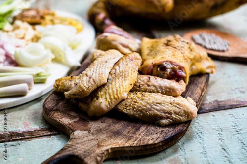 Pieces of fresh raw chicken parts on a cutting board on wooden old plank or table with blur Thai herbs and whole chicken. Chicken with Thai herbs prepared for cooking soup.