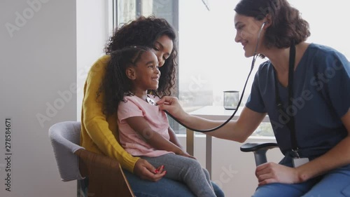 Female Pediatrician Wearing Scrubs Listening To Girls Chest With Stethoscope In Hospital Office