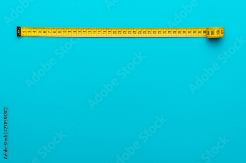 Top view of yellow soft measuring tape. Minimalist flat lay image of tape measure with metric scale over turquoise blue background with copy space.