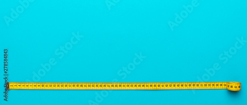 Top view of yellow soft measuring tape. Minimalist flat lay image of tape measure with metric scale over turquoise blue background. Panoramic orientation photo of tape measure with copy space.