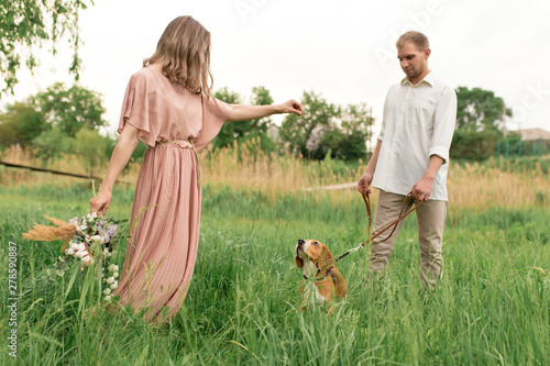 Young loving couple having fun and running on the green grass on the lawn with their beloved domestic dog breed Beagle and a bouquet of wildflowers