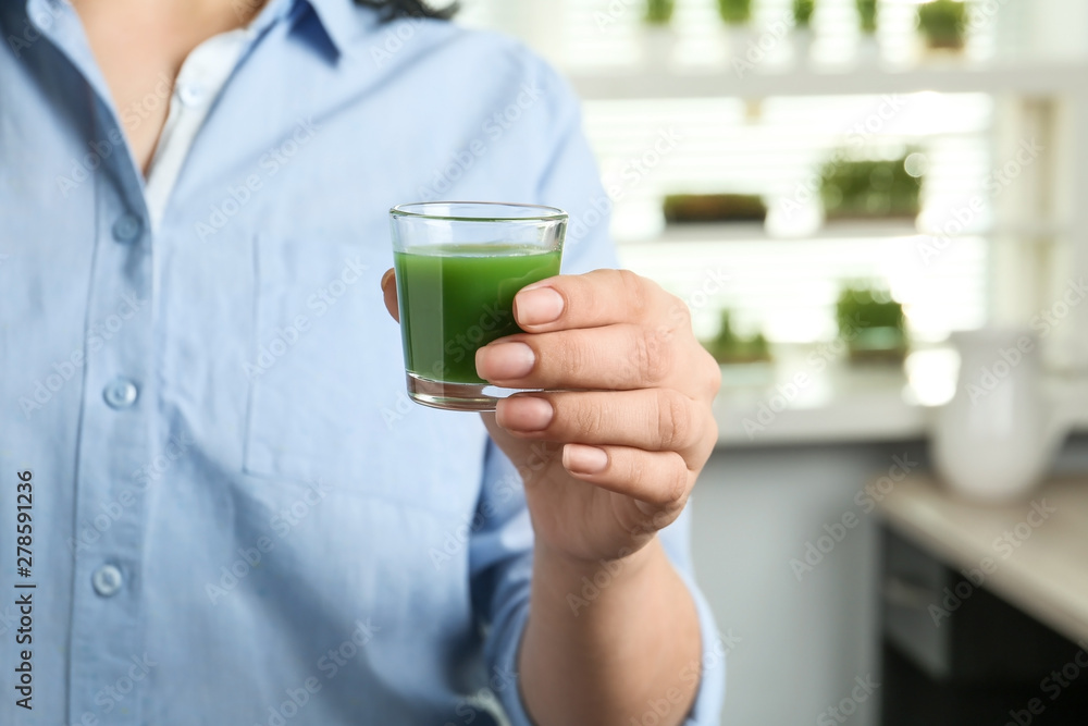 Woman holding shot glass of wheat grass juice indoors, closeup Woman holding shot glass of wheat grass juice indoors, closeup