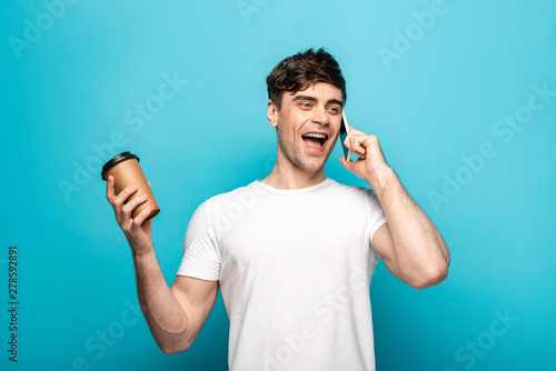 cheerful young man talking on smartphone and holding coffee to go on blue background © LIGHTFIELD STUDIOS