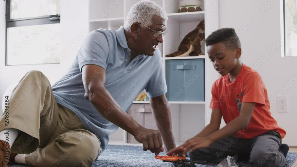 Grandfather With Grandson Sitting On Rug At Home Building Model Helicopter Together Grandfather With Grandson Sitting On Rug At Home Building Model Helicopter Together
