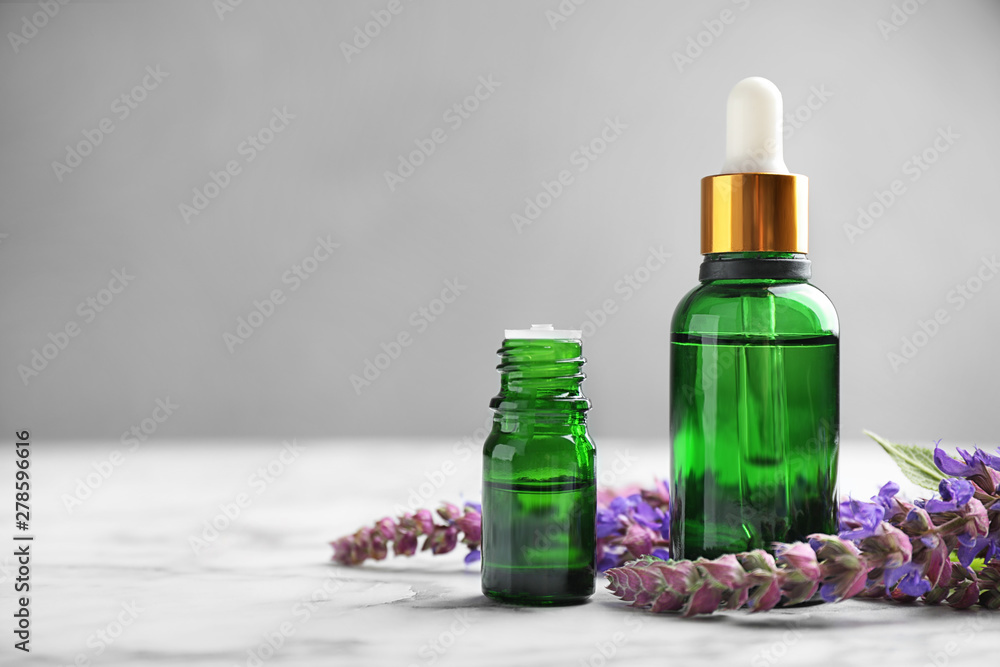 Bottles of sage essential oil and flowers on marble table, space for text