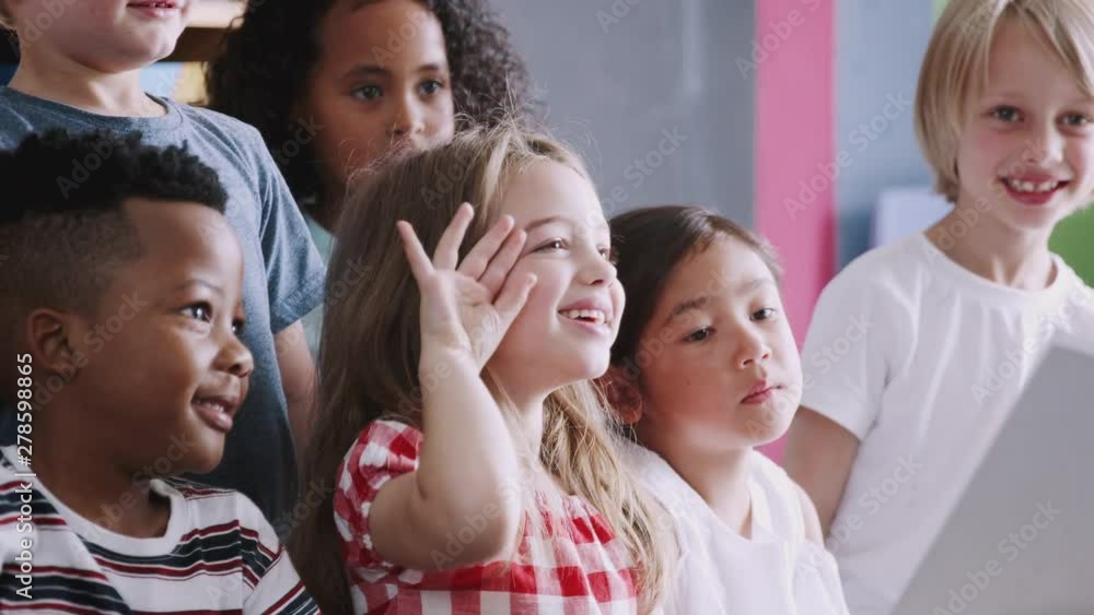 Elementary Pupils Raising Hands To Answer Question As Female Teacher Reads Story In Classroom Elementary Pupils Raising Hands To Answer Question As Female Teacher Reads Story In Classroom
