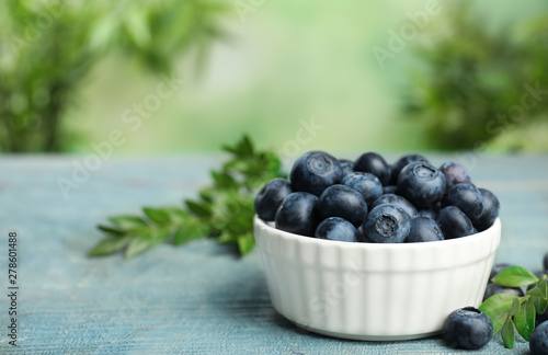 Bowl of tasty blueberries and leaves on wooden table against blurred green background, space for text