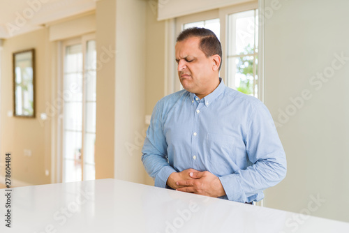 Middle age man sitting at home with hand on stomach because nausea, painful disease feeling unwell. Ache concept.