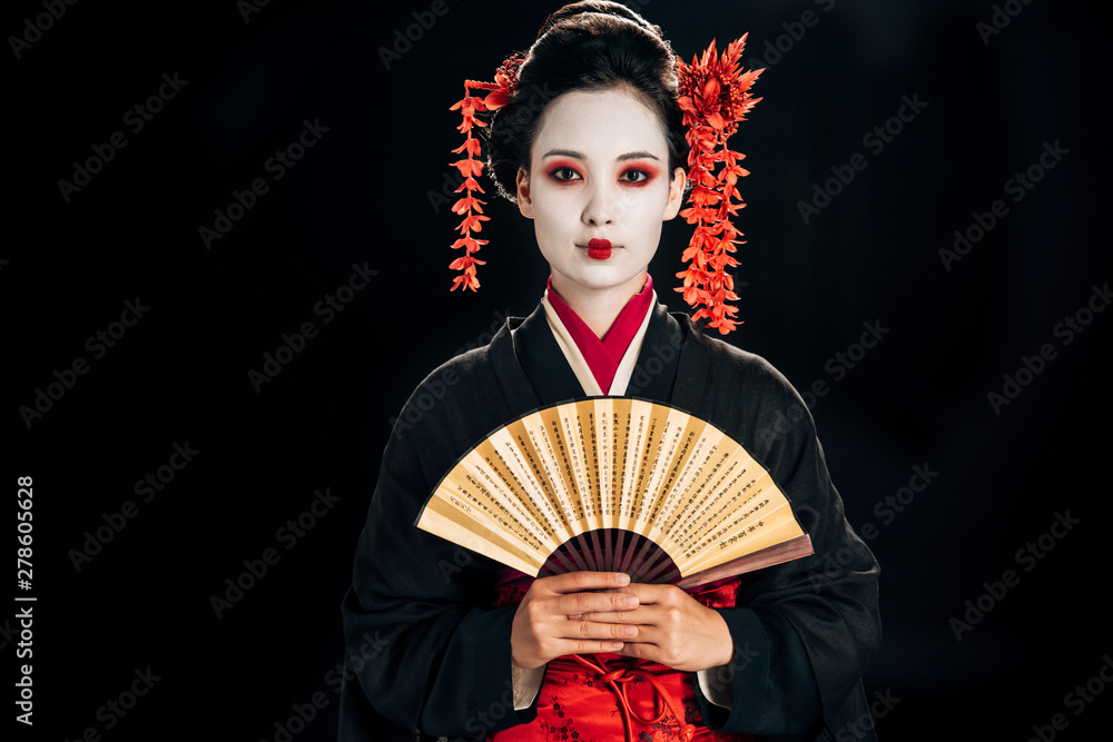 Obraz premium geisha in black kimono with red flowers in hair holding traditional asian hand fan isolated on black