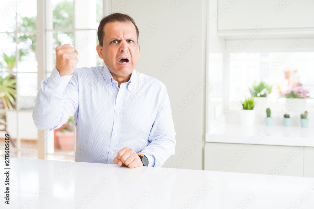 Middle age man sitting at home angry and mad raising fist frustrated and furious while shouting with anger. Rage and aggressive concept.