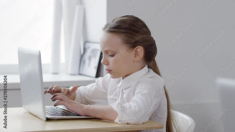 Chest-up shot of 9-year-old Caucasian schoolgirl, wearing white blouse, sitting at desk in computer class at school, working on assignment on laptop, typing on keyboard, looking down and up at screen Chest-up shot of 9-year-old Caucasian schoolgirl, wearing white blouse, sitting at desk in computer class at school, working on assignment on laptop, typing on keyboard, looking down and up at screen
