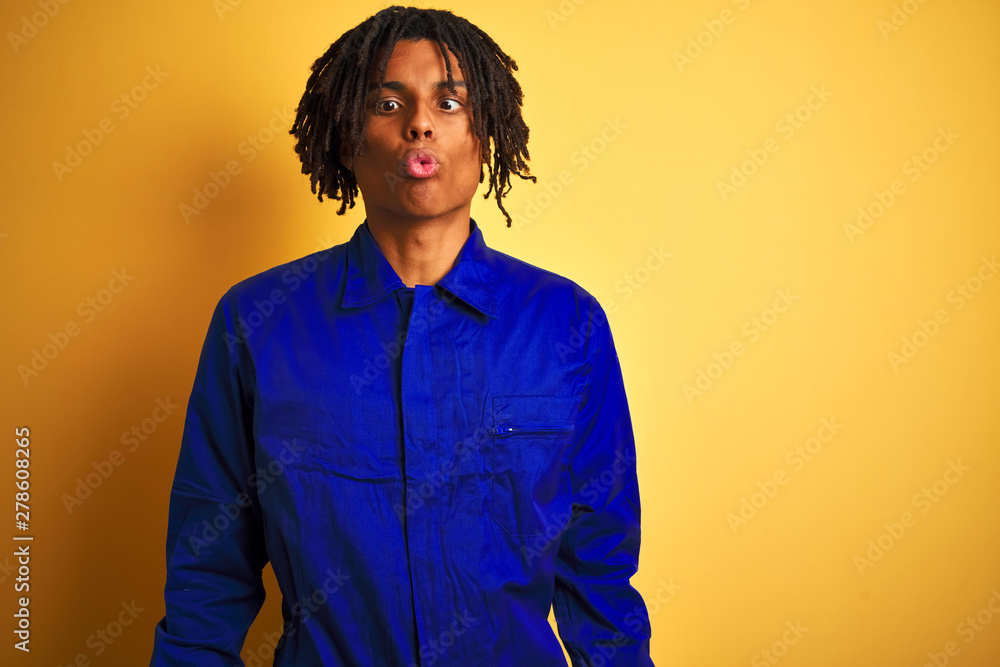 Obraz premium Afro worker man with dreadlocks wearing mechanic uniform over isolated yellow background making fish face with lips, crazy and comical gesture. Funny expression.