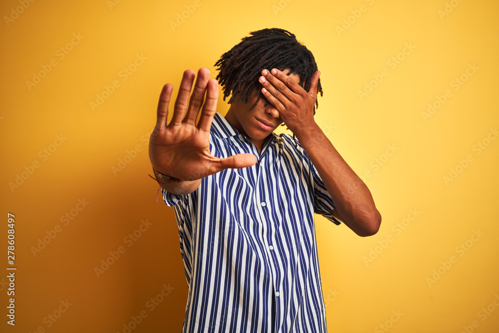 Afro man with dreadlocks wearing casual striped t-shirt over isolated yellow background covering eyes with hands and doing stop gesture with sad and fear expression. Embarrassed and negative concept. Afro man with dreadlocks wearing casual striped t-shirt over isolated yellow background covering eyes with hands and doing stop gesture with sad and fear expression. Embarrassed and negative concept.