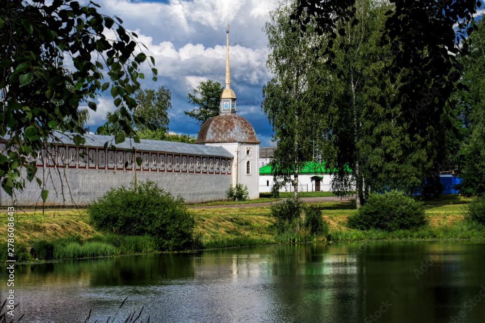 Medieval tower and fencing of  Tikhvinsky Monastery built in 16th century with a reflection in the pond surrounded by trees on a sunny summer day