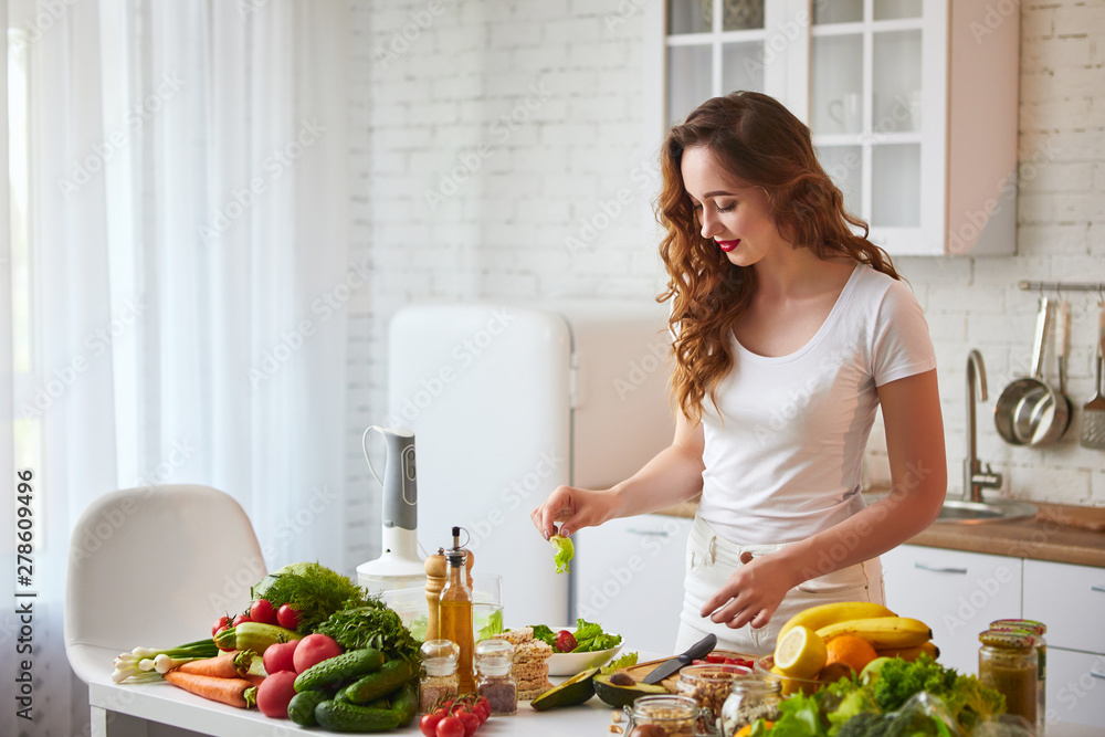 Young happy woman preparing tasty salad in the beautiful kitchen with green fresh ingredients indoors. Healthy food and Dieting concept. Loosing Weight Young happy woman preparing tasty salad in the beautiful kitchen with green fresh ingredients indoors. Healthy food and Dieting concept. Loosing Weight