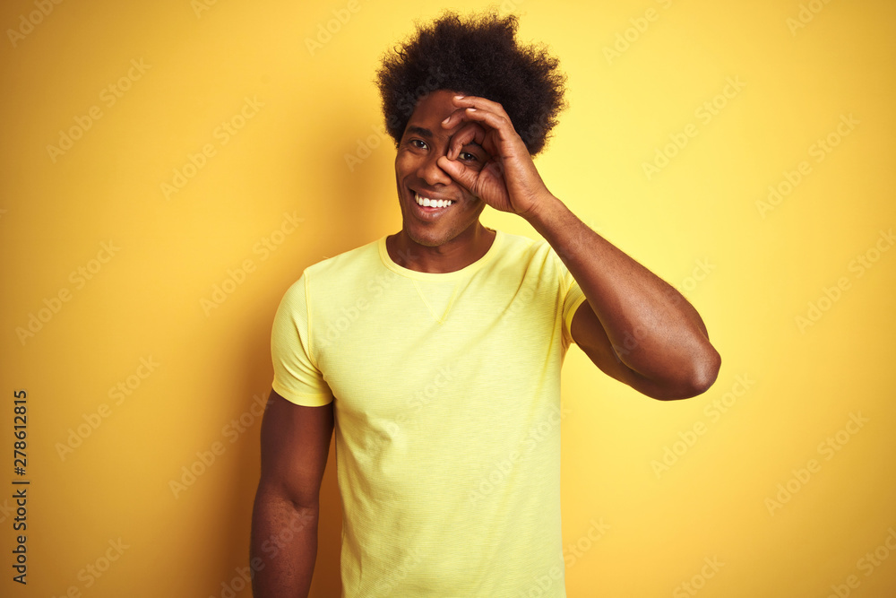 African american man with afro hair wearing t-shirt standing over isolated yellow background doing ok gesture with hand smiling, eye looking through fingers with happy face. African american man with afro hair wearing t-shirt standing over isolated yellow background doing ok gesture with hand smiling, eye looking through fingers with happy face.