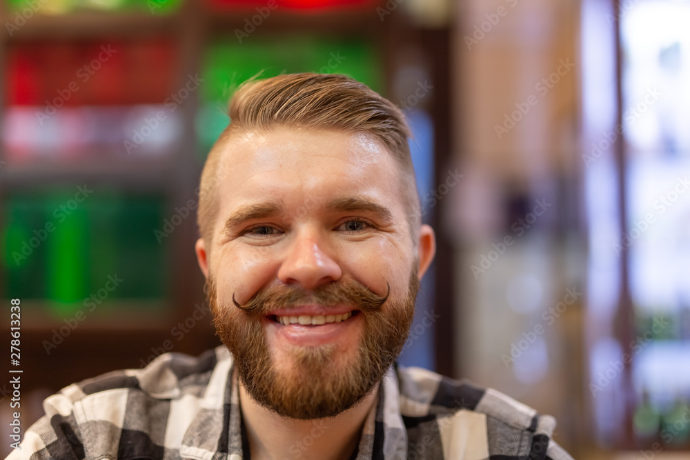 Cute positive young hipster guy with a mustache and beard smiling posing in a cafe. The concept of funny cheerful men. Cute positive young hipster guy with a mustache and beard smiling posing in a cafe. The concept of funny cheerful men.