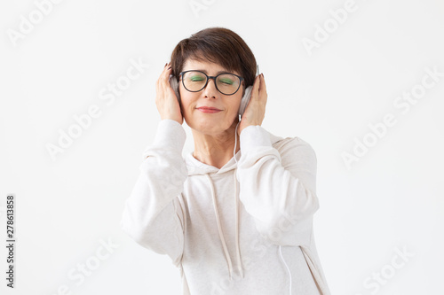 Pretty keen middle-aged woman in glasses and a white sweater listens to her favorite music with headphones on a white background with copy space. Online radio and music subscription concept.