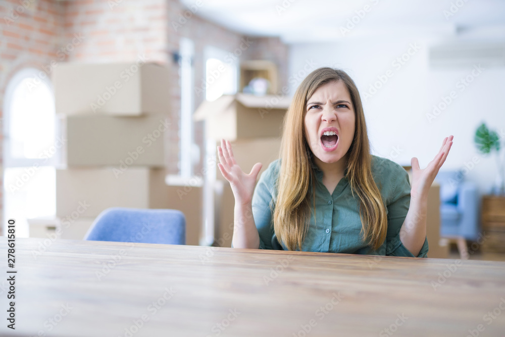 Young woman sitting on the table with cardboard boxes behind her moving to new home crazy and mad shouting and yelling with aggressive expression and arms raised. Frustration concept. Young woman sitting on the table with cardboard boxes behind her moving to new home crazy and mad shouting and yelling with aggressive expression and arms raised. Frustration concept.