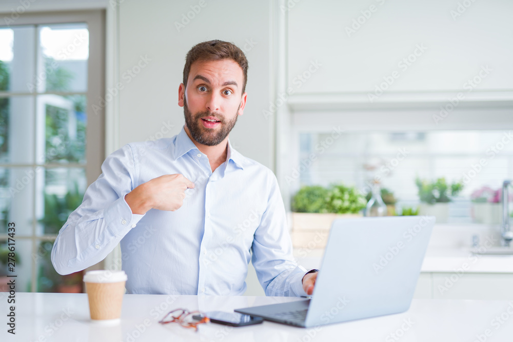 Handsome man working using computer laptop and drinking a cup of coffee with surprise face pointing finger to himself