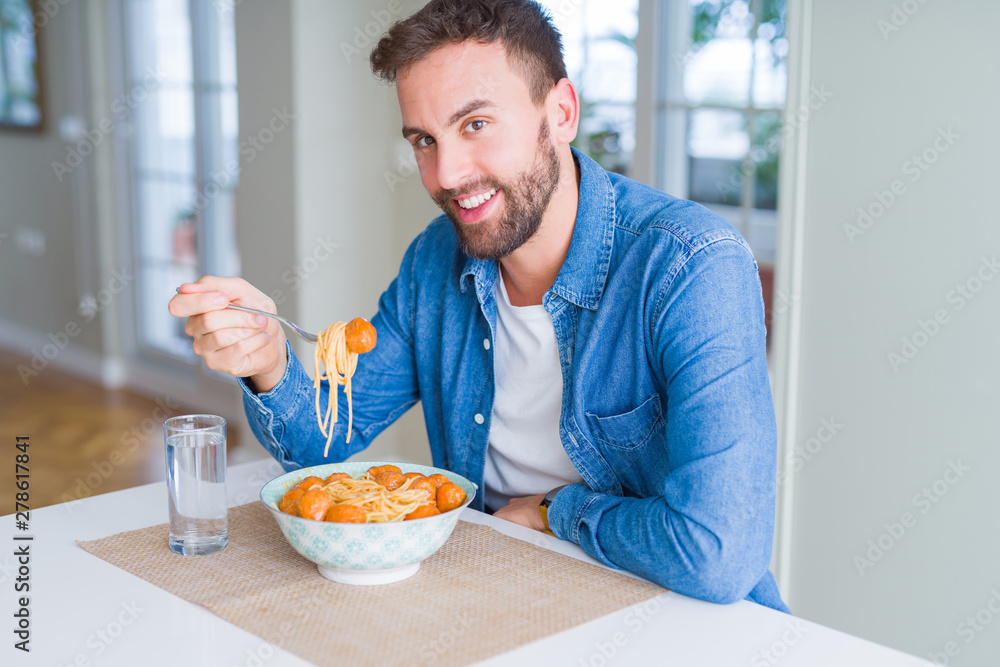 Handsome man eating pasta with meatballs and tomato sauce at home while smiling at the camera Handsome man eating pasta with meatballs and tomato sauce at home while smiling at the camera