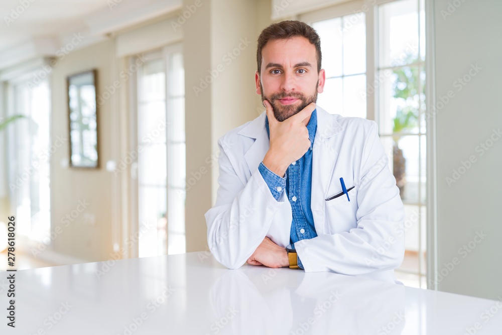 Handsome doctor man wearing medical coat at the clinic looking confident at the camera with smile with crossed arms and hand raised on chin. Thinking positive. Handsome doctor man wearing medical coat at the clinic looking confident at the camera with smile with crossed arms and hand raised on chin. Thinking positive.