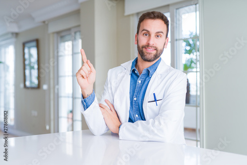 Handsome doctor man wearing medical coat at the clinic with a big smile on face, pointing with hand and finger to the side looking at the camera.